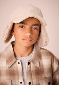 Beige fleece hat med øreklapper, kombineret med en ternet skjorte i brune og creme farver. Grå t-shirt under, glat tekstur og afslappet design.