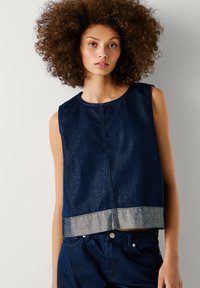 Tumesinise ilma varrukateta teksasülikond, millel on läikiv hõbeäär. Omab ümarat kaelust ja ruudukujulist lõiget, rõhutades sujuvat tekstuuri.