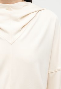 Κοντινή άποψη ενός ατόμου που φοράει ένα ελαφρύ, κρεμ φούτερ με κουκούλα, με χαλαρό, επικαλυπτόμενο λαιμόκοψη και μακριά μανίκια.