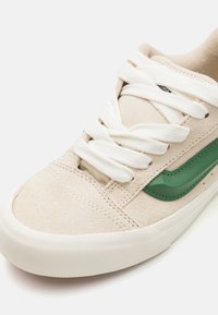 Beige suede sneaker with white laces, featuring a dark green logo accent. Round toe and textured rubber sole with a smooth finish.