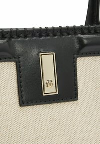 Borsa in canvas con corpo crema, dettagli in pelle nera e un particolare in metallo rettangolare color oro con design in rilievo.