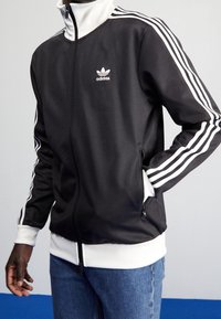 Svart polyester-träningsjacka med vita ränder på ärmarna, hög krage och dragkedja. Har en Adidas-logotyp på bröstet.