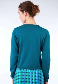 Türkiser, langärmeliger Pullover mit rundem Halsausschnitt, aus weichem Material. Tragen mit einem gemusterten blau-grünen Rock mit ovalen Formen.