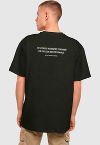 Sort bomulds t-shirt med løs pasform, der har hvid tekst på ryggen, som siger "DEN ULTIMATIVE MOTORSPORTSLEDSSAGER FOR PRÆCISION OG YDEEVNE."