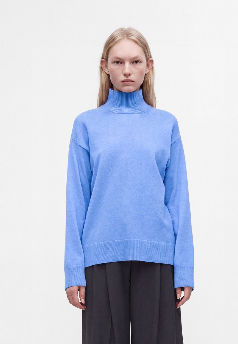Jeune personne aux longs cheveux blonds portant un pull à col roulé bleu clair et un pantalon ample foncé sur fond blanc uni.