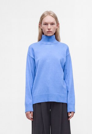 Jeune personne aux longs cheveux blonds portant un pull à col roulé bleu clair et un pantalon ample foncé sur fond blanc uni.