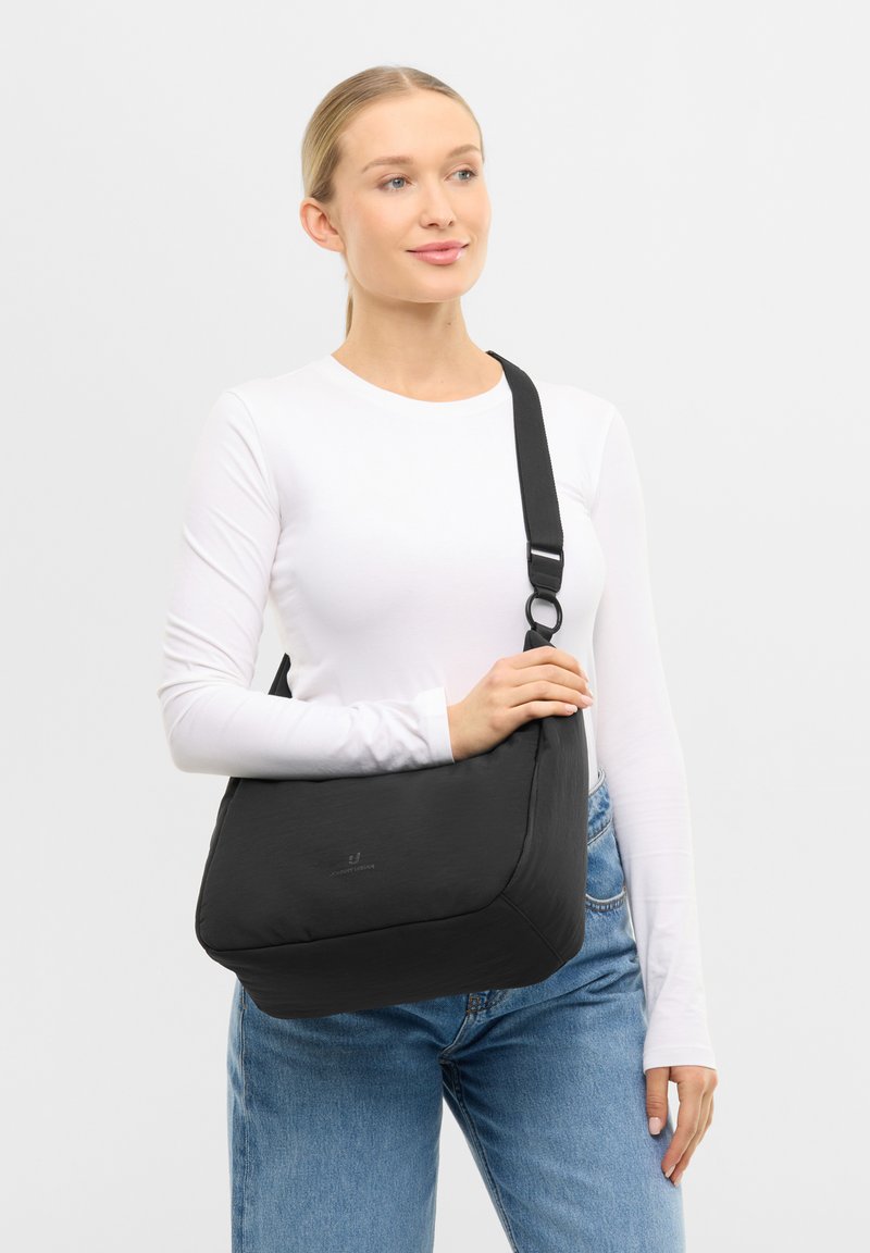 Femme portant une chemise blanche à manches longues et un jean, avec un grand sac bandoulière noir à sangle réglable sur l'épaule.