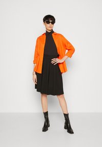 Camicia arancione a bottoni sopra un dolcevita nero, abbinata a una gonna plissettata nera e stivaletti neri. Gli accessori includono occhiali da sole e anelli.