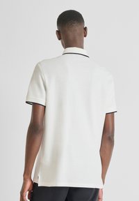 Polo bianco a maniche corte, con bordi neri su colletto e maniche, texture liscia e lati traforati. La vista mostra il retro della maglietta.