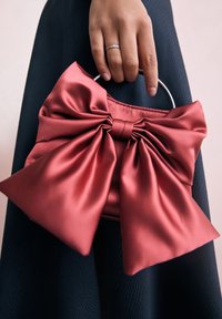 Rdeča torbica iz satena z velikim mašnim detajlom, okroglim kovinskim ročajem in teksturirano črno tkanino v ozadju. Roka drži torbico.