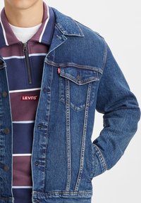 Chaqueta de mezclilla en azul medio con cuello puntiagudo clásico, tapeta de botones, bolsillo en el pecho y logo de Levi's. Llevada sobre una camisa de cremallera a rayas.