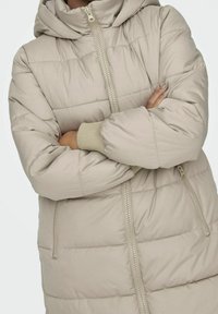 Beige gewatteerd jack met capuchon, voorzien van een ritssluiting en geribbelde manchetten. Het materiaal is glad met een licht glanzende textuur en gestikte panelen.