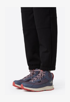 Chaussures de trail avec une tige en mesh bleu marine et violet, des accents rouges et une semelle en caoutchouc texturée, associées à un pantalon noir fuselé.