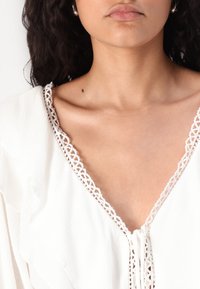 Blusa bianca con un bordo in pizzo decorativo lungo il scollo a V, texture morbida e vestibilità ampia con pieghe sottili sul tessuto.