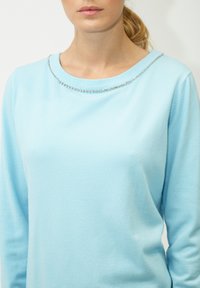 Hellblauer Pullover mit langen Ärmeln, der einen runden Halsausschnitt mit einem silbernen Perlenakzent entlang des Kragens hat. Weiche Textur.