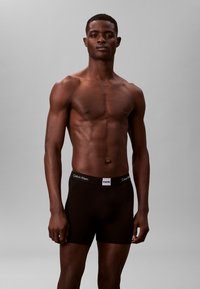 Czarne bokserki z bawełny z białym logo "Calvin Klein" na gumce, model stoi na jednolitym szarym tle.