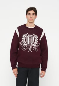 Bordo pletena majica s bijelim detaljima i Adidas logom s lovorovim listovima, okruglim izrezom i rebrastim manžetama i rubom.