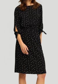 Robe noire à pois blancs, à manches trois-quarts, taille élastique et accents noués aux bords des manches. Texture de tissu lisse.