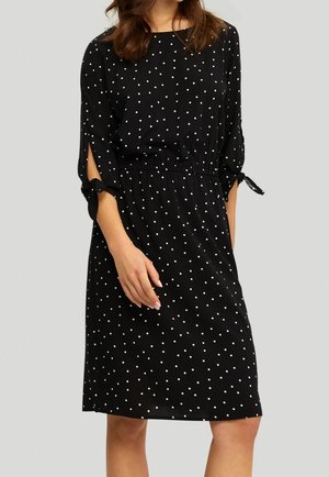 Robe noire à pois blancs, à manches trois-quarts, taille élastique et accents noués aux bords des manches. Texture de tissu lisse.