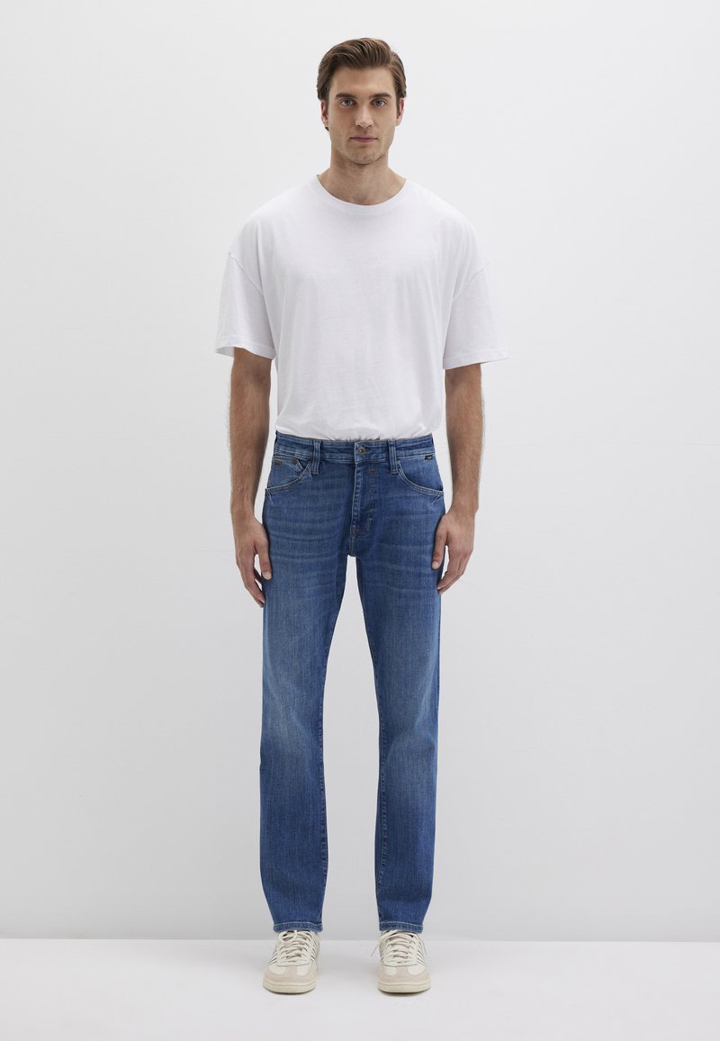 Leichtes weißes Baumwoll-T-Shirt, kombiniert mit blauen Jeans. Die Jeans haben einen geraden Schnitt, Fronttaschen und eine dezente Waschung. Schlichte beige Sneakers.