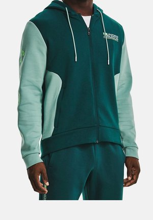 Homme portant un sweat à capuche zippé Under Armour vert et vert clair avec un pantalon de survêtement vert assorti, debout avec les mains détendues le long du corps.