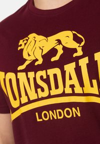 Burgundi színű pamut póló, sárga oroszlán grafikával és vastag sárga "LONSDALE LONDON" felirattal a mellkasán.