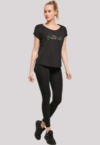 Juodas trumpas rankovėmis marškinėliai su "Disney Princess" grafika spalvingu tekstu, derinami su juodais leggingais ir juodais sportiniais batais.