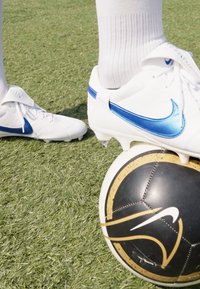 Valged Nike jalgpalliklipid siniste aktsentidega rohelisel murul, paigutatud musta ja kuldse Nike jalgpalliga. Sokid on valged ja tekstuuriga.