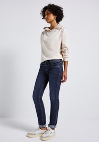 Beige hoodie med en cowl neck, tillsammans med mörkblå slim-fit jeans. Outfiten kompletteras av vita sneakers med beige detaljer.