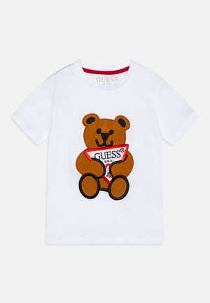 T-shirt in cotone bianco con un orsetto di peluche ricamato di colore marrone che tiene un banner rosso con la scritta "GUESS U.S.A." in nero. Maniche corte.