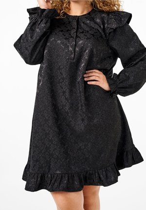 Zwarte jurk met lange mouwen met een subtiel bloemmotief, gerimpelde schouders en zoom, gedragen door een persoon die poseert met één hand op de heup.