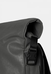 Grijze rugzak met roll-top, gemaakt van glad synthetisch materiaal, voorzien van een zwarte gesp-strapsluiting en zichtbare stikdetails.