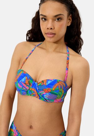 Mujer con un top de bikini de estampado tropical colorido con tirantes halter, mostrando el torso superior y un fondo neutro.