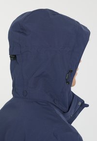 Mörkblå huva jacka gjord av texturerat tyg. Har justerbara snören på huvan, tryckknappar och en hög krage för extra skydd.