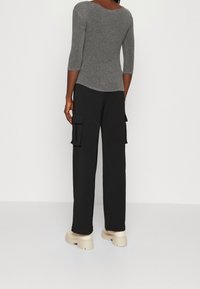 Kvinna iklädd en åtsittande grå topp med trekvartslånga ärmar, svarta cargobyxor med stora fickor och beige klumpiga platåskor som står vänd bort.