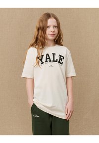 Izlīdzena krēmkrāsas T-krekls ar īsām piedurknēm, uz kura ir biezām melnām burtiem uzrakstīts "YALE". Apvienots ar tumši zaļām biksēm, demonstrējot relaksētu piegriezumu.