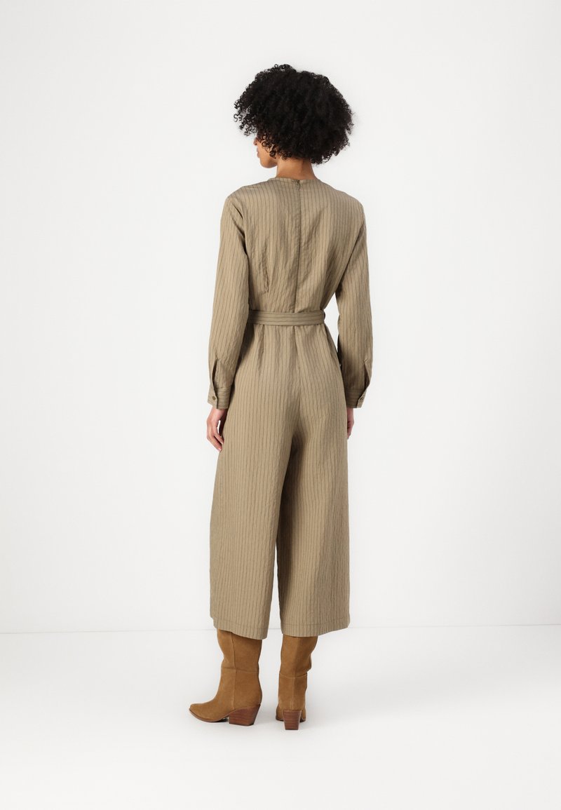 Olivgrüner, gerippter Jumpsuit mit langen Ärmeln, tailliertem Gürtel und weiten Hosenbeinen, kombiniert mit kniehohen, braunen Suede-Stiefeln.