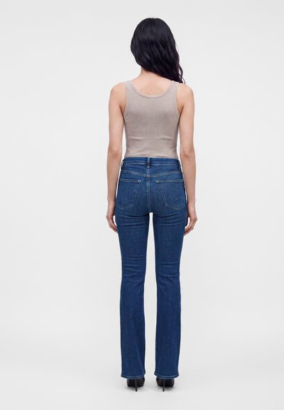 Femme aux longs cheveux noirs debout de dos, portant un top beige sans manches, un jean bleu et des chaussures à talons noires sur un fond uni.