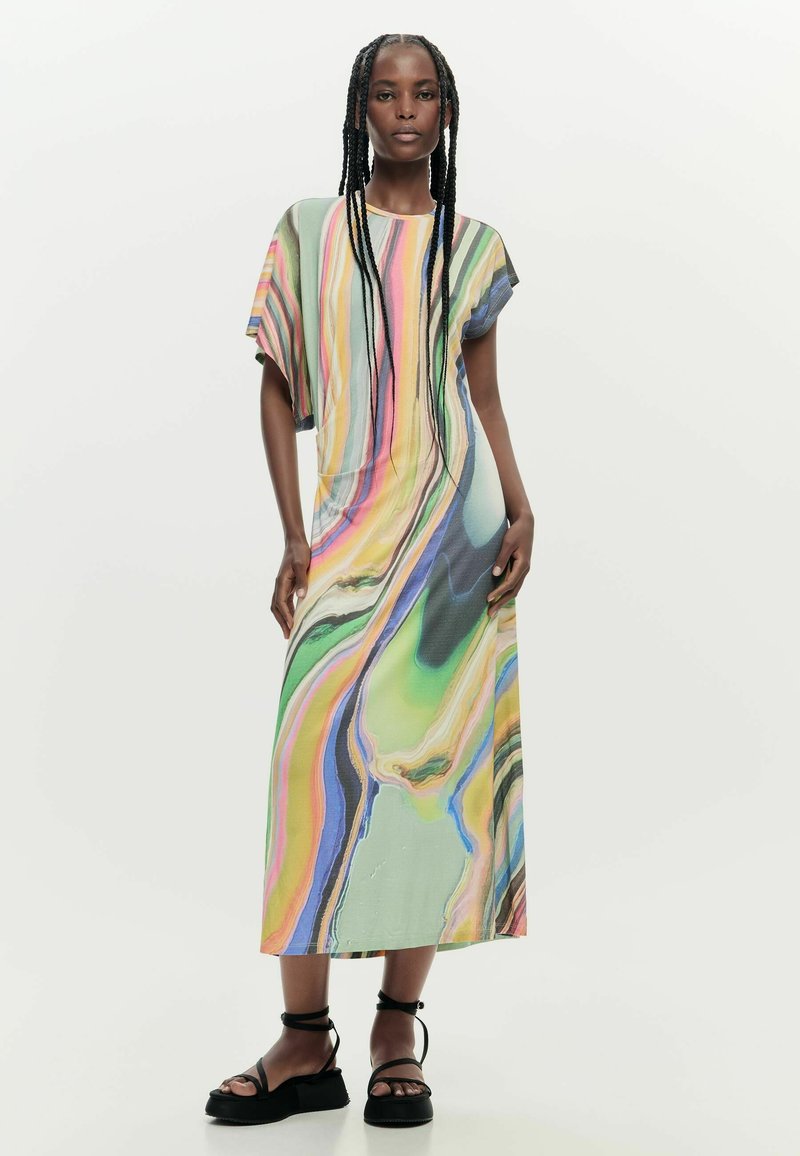 Nő egy fehér háttér előtt áll, színes, márványmintás midi ruhát visel rövid ujjakkal és fekete bokapántos platform szandált.