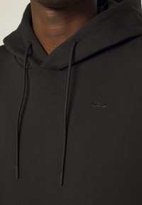 Černá bavlněná mikina s klokaní kapsou, s decentním vyšitým logem a tkaničkami. Hladká textura a volný střih.