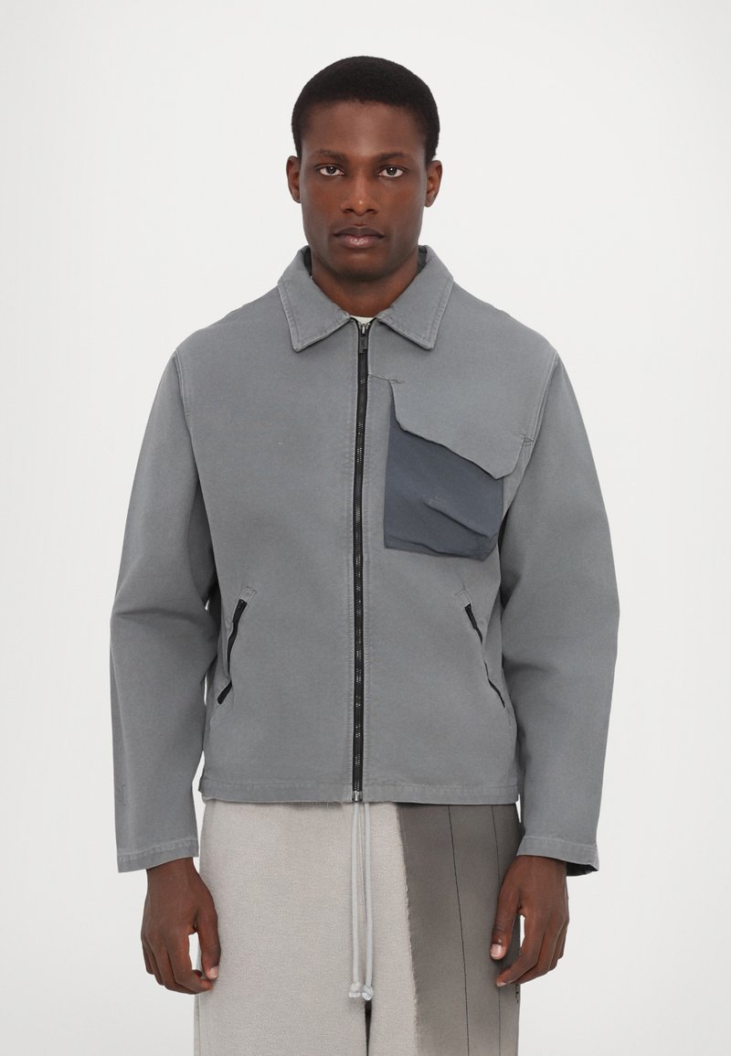 Veste grise en tissu durable, avec une fermeture éclair à l'avant, une grande poche poitrine et des poches latérales. Texture lisse avec un design structuré.