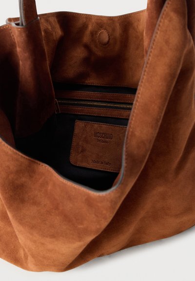 Καφέ τσάντα από σουέντ με ανοιχτό πάνω μέρος, εσωτερική τσέπη με φερμουάρ και ορατή δερμάτινη ετικέτα Moschino Milano με την ένδειξη Made in Italy.