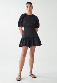 Czarna sukienka z gładkiego materiału, z krótkimi puffowanymi rękawami, marszczonym pasem i falbaniastym dołem. Noszona z czarnymi sandałami.