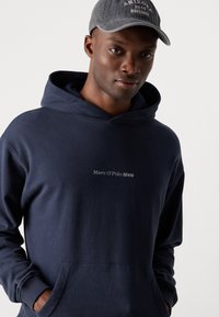 Hanorac cu glugă de culoare bleumarin, confecționat din bumbac, având o buzunar tip cangur și un logo alb "Marc O'Polo DENIM" pe piept.