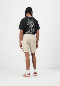 Zwarte katoenen t-shirt met een gedetailleerde graphic op de achterkant, gecombineerd met beige corduroy shorts en rood-gestreepte sneakers.
