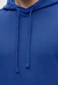 Gros plan d'une personne portant un sweat à capuche bleu avec des cordons épais et un logo "HUGO" subtilement embossé sur la poitrine.
