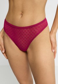 Transparante magenta string met een gestippeld patroon, ingewikkelde scalloped randen en een gladde textuur, gepositioneerd op het lichaam van een model.