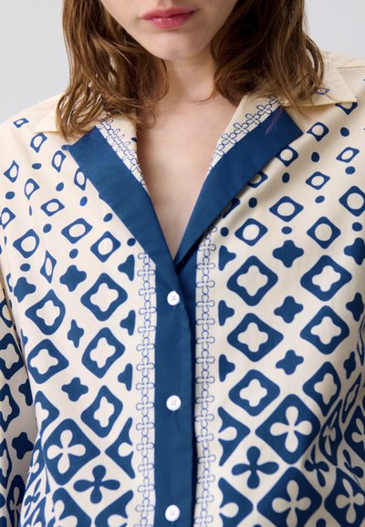 Camicetta a maniche lunghe color crema con motivi geometrici blu, dotata di colletto blu e chiusura con bottoni. Tessuto morbido con una finitura elegante.