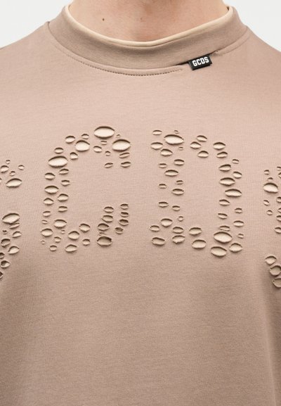Μπεζ κοντομάνικο μπλουζάκι με ανάγλυφα σχέδια σε σχήμα φυσαλίδων που σχηματίζουν τα γράμματα "GCDS" κοντά στο πάνω μέρος του στήθους και μια μικρή μαύρη ετικέτα της μάρκας στο γιακά.