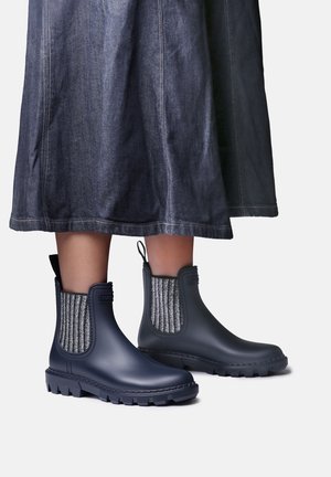 Personne portant des bottines de pluie bleu foncé avec des panneaux en tissu extensible gris et une jupe en jean foncé.